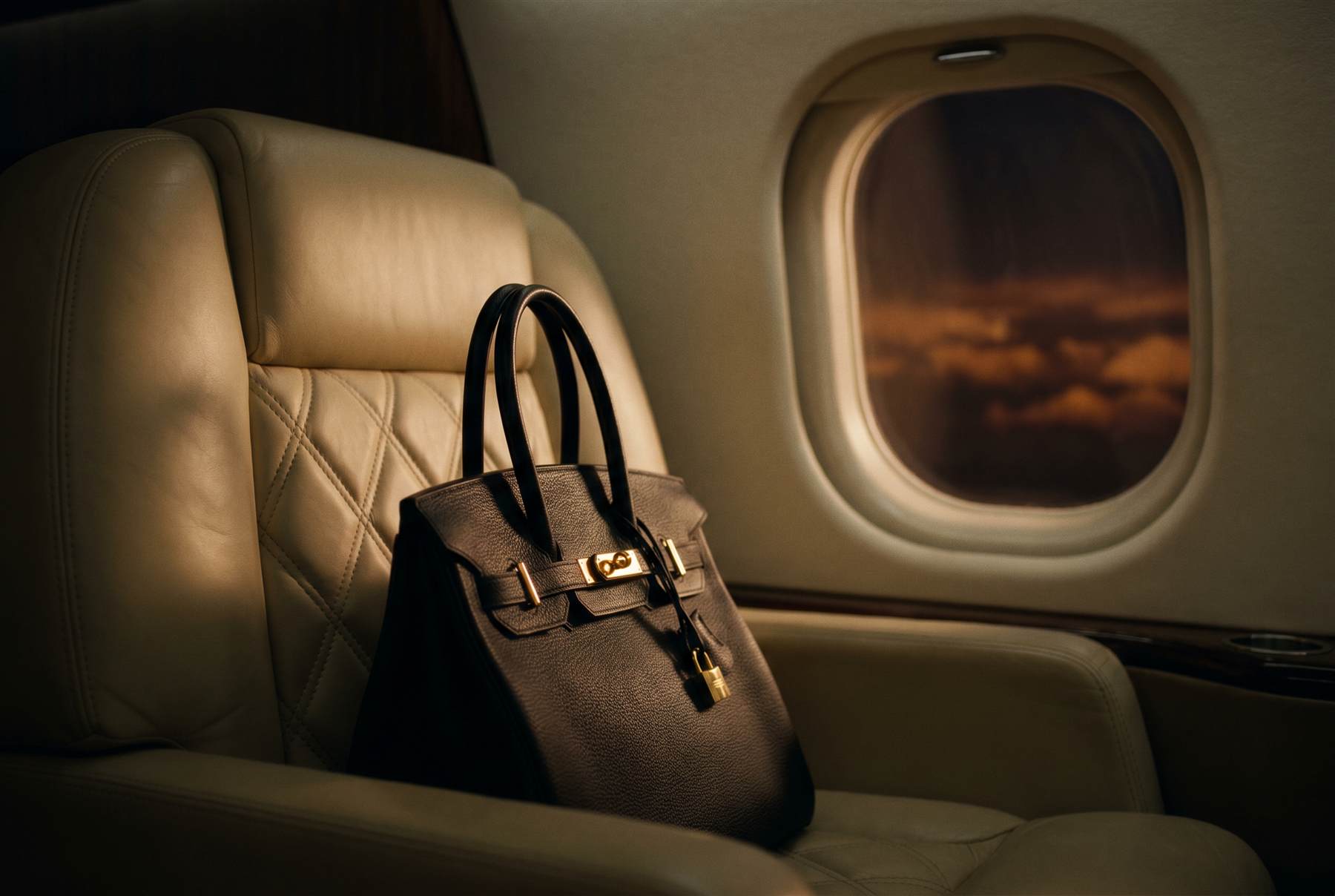 Black leather handbag with gold hardware on a cream leather jet seat beside a porthole window